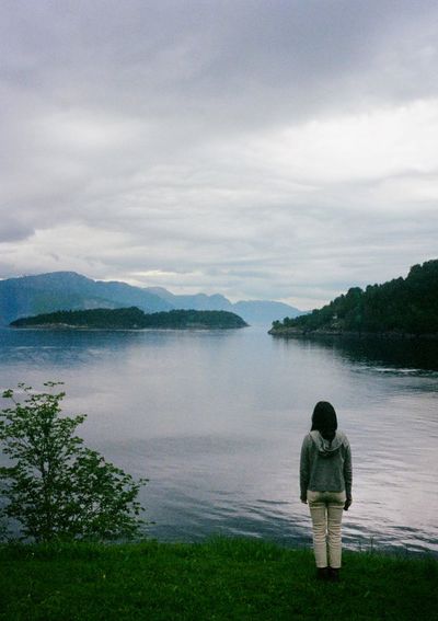 A woman with her back turned to the camera looks out across a fjord under grey, cloudy skies.