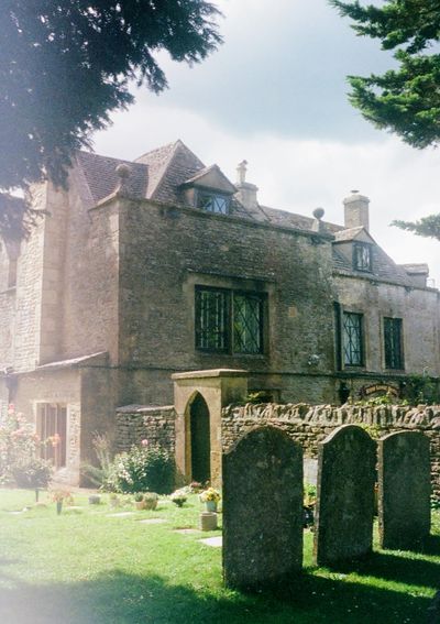 Three gravestones are in the foreground of this picture, with lush grass and a stone wall leading separating them from a house.