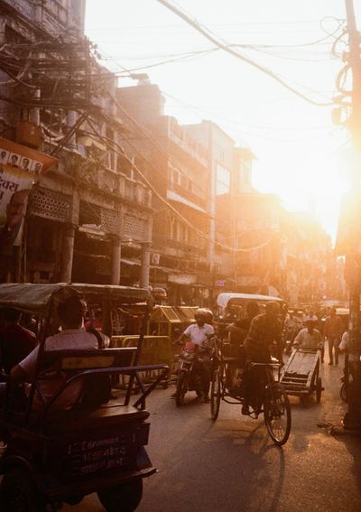 Rickshaws and lorries move in narrow, crowded streets in between old buildings. The evening sun shines bright into the shot.