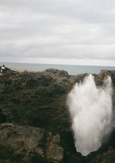 Against a rocky backdrop, a splash of ocean spray rises out of a blow hole. The sea is in the background.