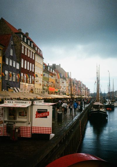 Boats line the banks of Nyhavn in Copenhagen. Besides them, tourists walk past restaurants along the shoreline.