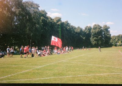 A crowd of people sit by the sideline of a playing field, with a large Tongan flag in the center of their group.