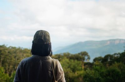 A female wearing hiking clothes has her back to the camera. She is looking out over green trees that extend out over hills in the background.