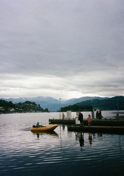 A man stands with a girl on a wooden pier, while a boy carefully navigates a small, yellow, inflatable boat towards them.