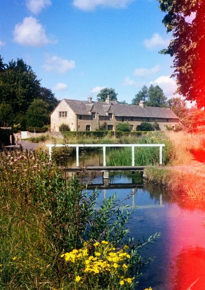 A stream flows through the town of Upper Slaughter in The Cotswolds. Stone brick houses line the roads to either side.