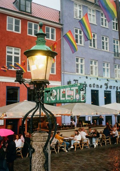 A sign for a public toilet is plastered with stickers. In the background a row of pride flags blow in the breeze.