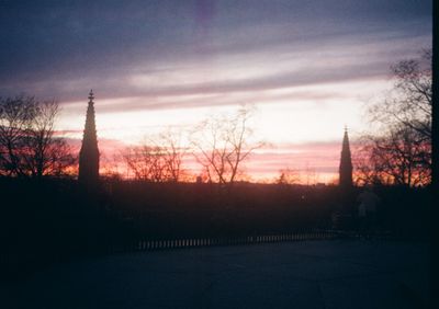 Church spires stick up above the skyline that's also dotted with trees. The setting sun in the background casts a orange and purple hue.