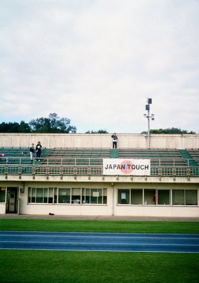 Three people stand separated in an otherwise empty grandstand. A sign in front of them reads 