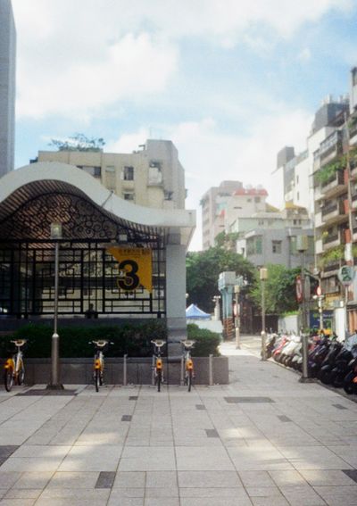 A row of public rental bikes stand at docks along the wall of a metro exit. The exit sign is number '3'.