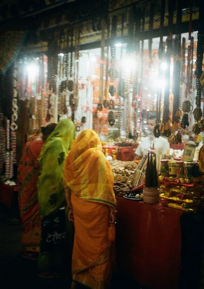 Three women in bright orange and green saries look at jewellery and bracelets in a brightly lit stall.