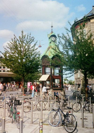 A small coffee house is in an old looking hectagonal building. It is in the middle of a bicycle parking lot, with bikes littered around it. Customers sit at the open windows facing the barista on the inside.