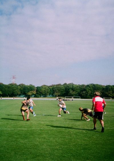 Men playing a game of Touch Football, with a person in a light cream shirt attacking the scoreline while a player in a dark jersey has slipped over while defending.