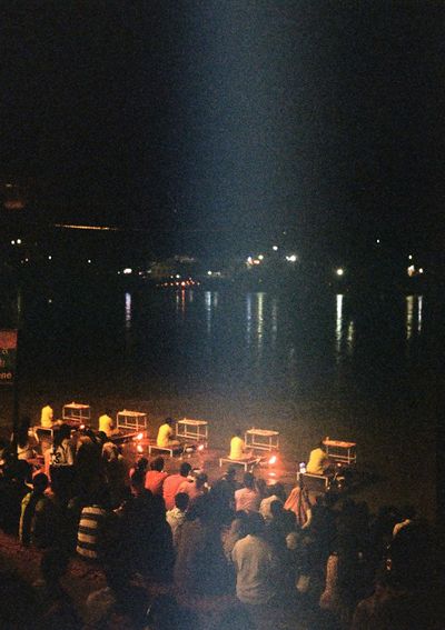 Five people sit on a platform at the edge of a river at night. In front of them 5 fires a lit. Behind them a large crowd of people watch on.