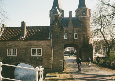 A man rides towards the camera, while two other play with a remote controlled car walking away from the photographer. All the men are walking under the arch of an old brick building.