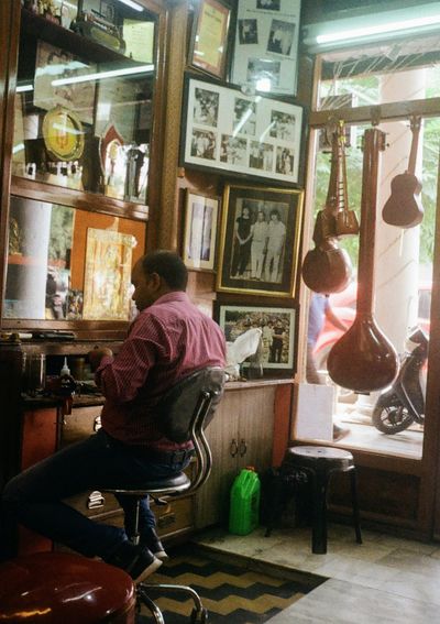 A man sits at a workbench in a shop. Sitars and other stringed instruments hang in the window behind him.