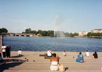 People sit facing a lake under the summer sun & clear skies. A gyser in the middle of the lake shoots water into the sky, creating a small rainbow.