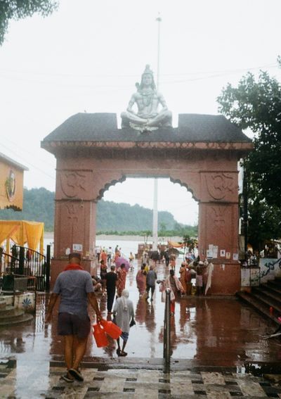 People walk away from the camera, under an archway topped with a statue of a deity. They are heading towards a river that is just visible in the background.