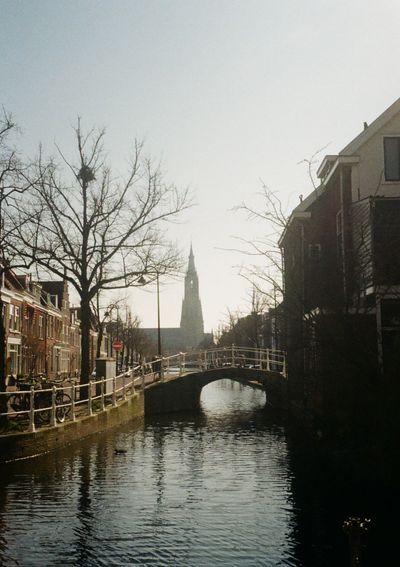 Looking down a canal lined by brick buildings. A bridge is in the middle of the frame, with a church tower in the distance background. The whole scene is filtered through late afternoon light.