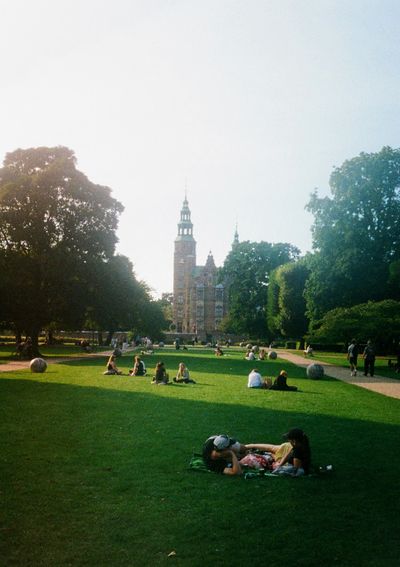 A couple lays on the grass in a park surrounded by trees. A palace is centered in the back of the photo.