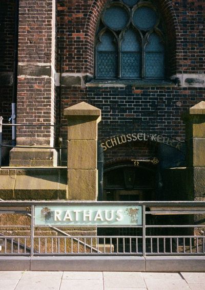 A sign saying 'Rathaus' is in the foreground of the image, place on a steel railing. In the background is an old brick building with a sign saying 'Schlussel Keller'.