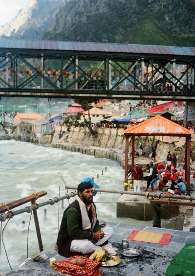 A holy man sits besides the empty plates of a finished ritual. A river flows under a bridge behind him.