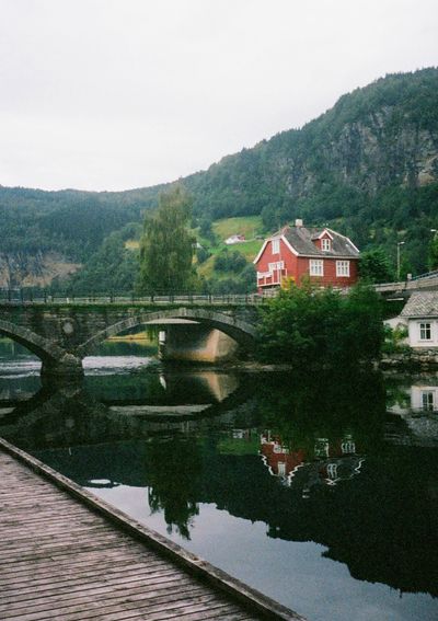 A house and bridge is reflected in the still waters of a fjord. The sky is overcast, and the water is calm.