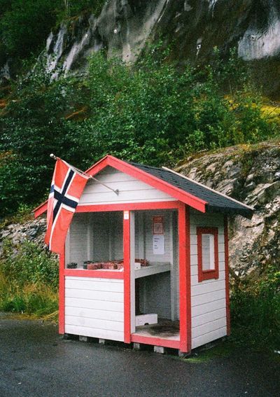 A white hut with red trimming and a black roof sits in a pulloff area by a road. It has a Norwegian flag flying from its roof. Inside are a collection of fruits and berries for sale. There's no person at the stall, it's based on an honesty system.