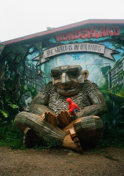 A boy climbs into the hands of a giant wooden troll statue. A mural painted behind it says 'Wonderland - The World is in our hands'.