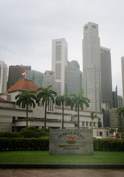 A stone sign saying Parliament sits in front of a neat lawn. City skyscrapers line the background.