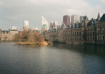 A view across a lake. An island in the middle sits in front of a very old brown brick building. Glass towers rise up in the background.