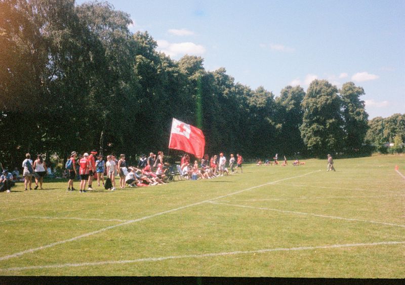 A crowd of people sit by the sideline of a playing field, with a large Tongan flag in the center of their group.