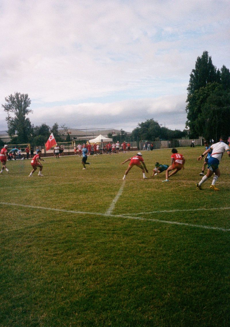 A man wearing a blue Japanese shirt dives low for the scoreline, holding a rugby ball in one hand. Two players wearing red Tonga shirts reach down to try and touch him.