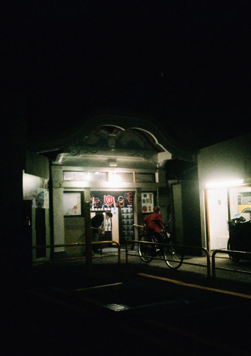 A lady pushes a bike down a ramp, while another ducks under a banner at the entrance of a public bath.