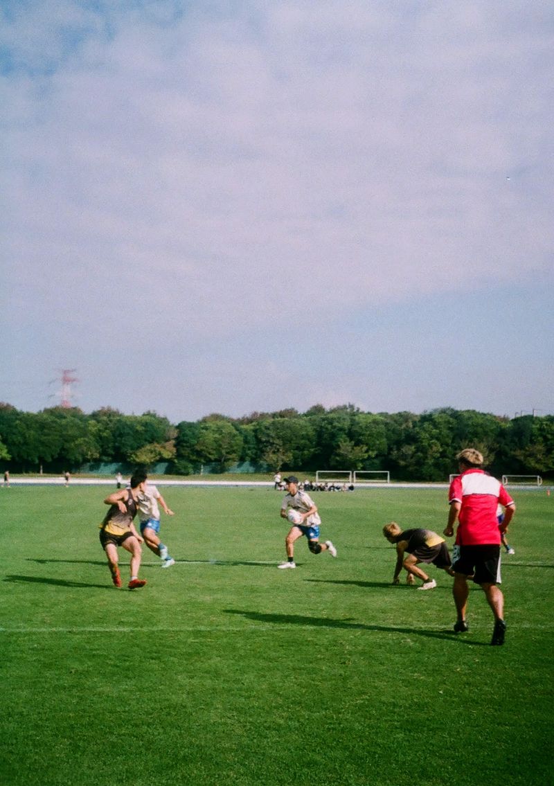 Men playing a game of Touch Football, with a person in a light cream shirt attacking the scoreline while a player in a dark jersey has slipped over while defending.