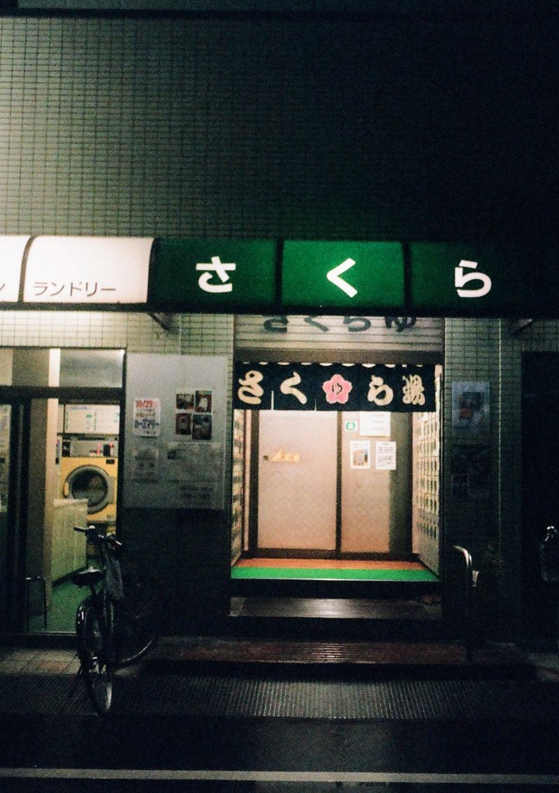 The entrance of a Japanese public bath is lit up at night. The roofing covering the entrance is green in colour, with some Japanese characters written across it.