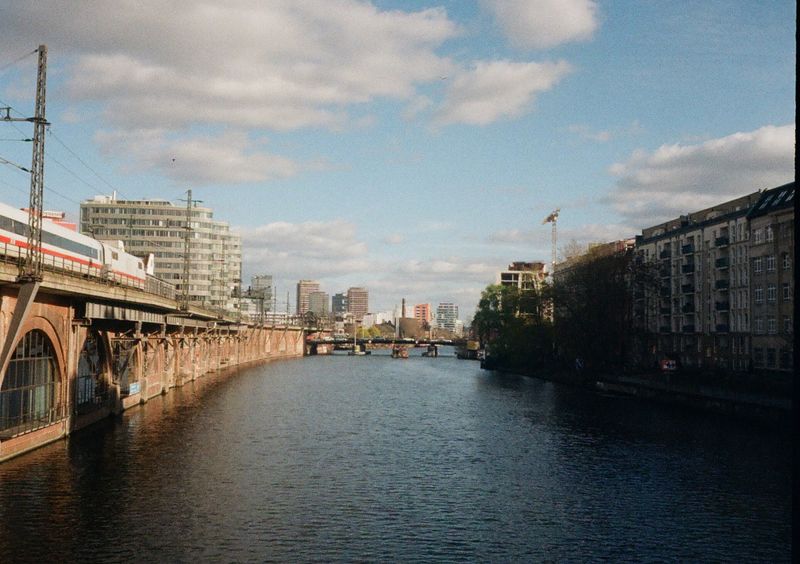 Looking down a river under a blue, clouded sky. On the left, a white train sits atop a brick embankment. On the right of the image are apartment buildings in the shade.