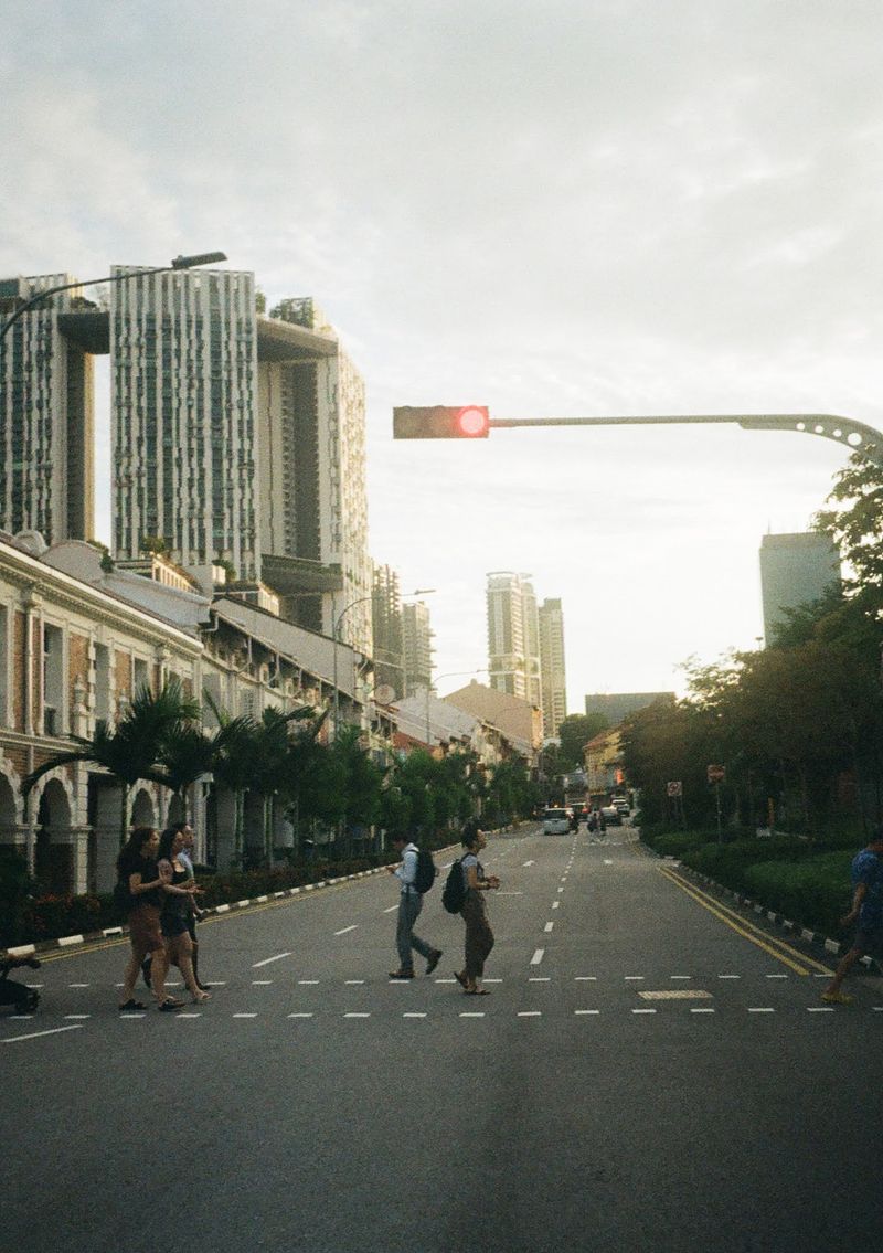 A photo taken while crossing the road, looking down the road towars highrise residential apartments in the distance. Pedestrians cross the road in the foreground, with older Chinese style houses on the left of the road.