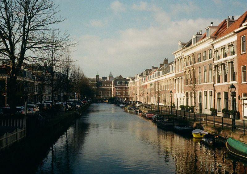 A look down a canal, with half the frame in sunlight and the other in shade. Boats line the sunny bank on the right with classy brick houses along the road next to the canal.