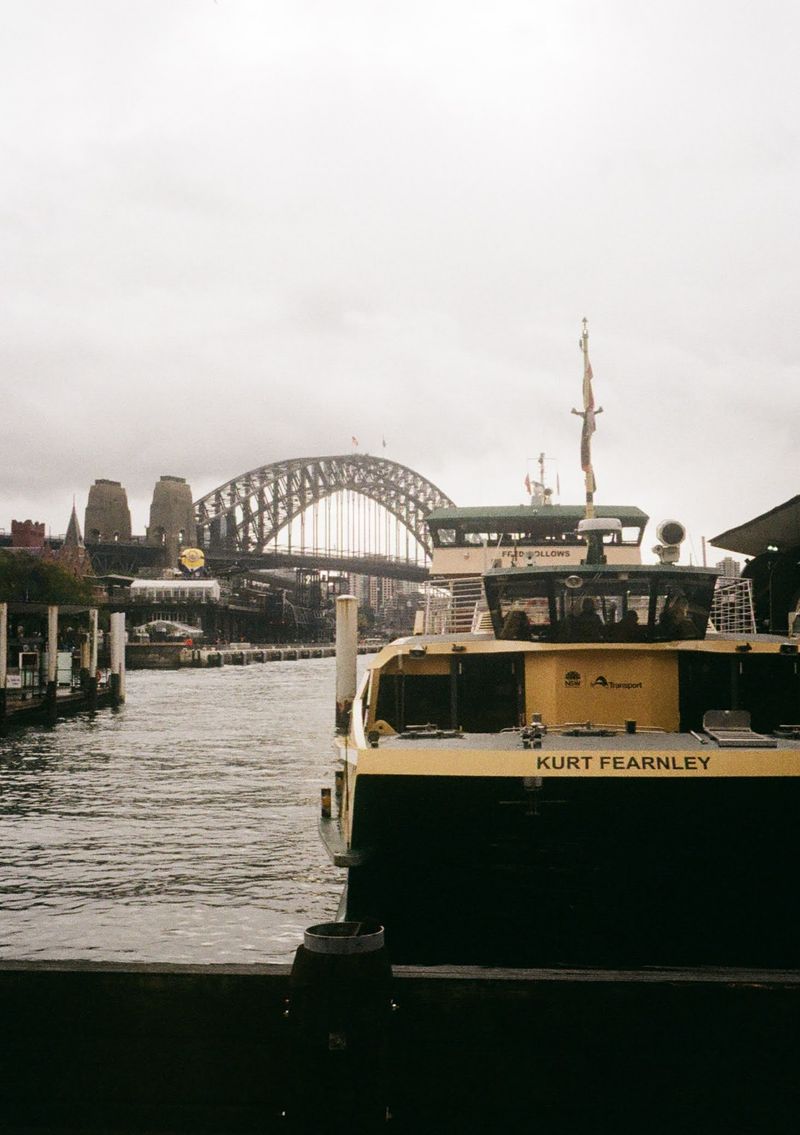 An old yellow and green Sydney Ferry is sitting by the dock with the Sydney Harbour Bridge in the background.