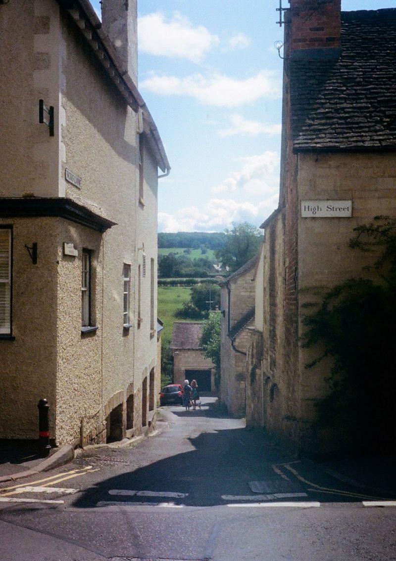 Two ladies walking down a lane that falls away out of the shot. Its entrance is between two old brick buildings, one which has a street sign saying 'High Street'.