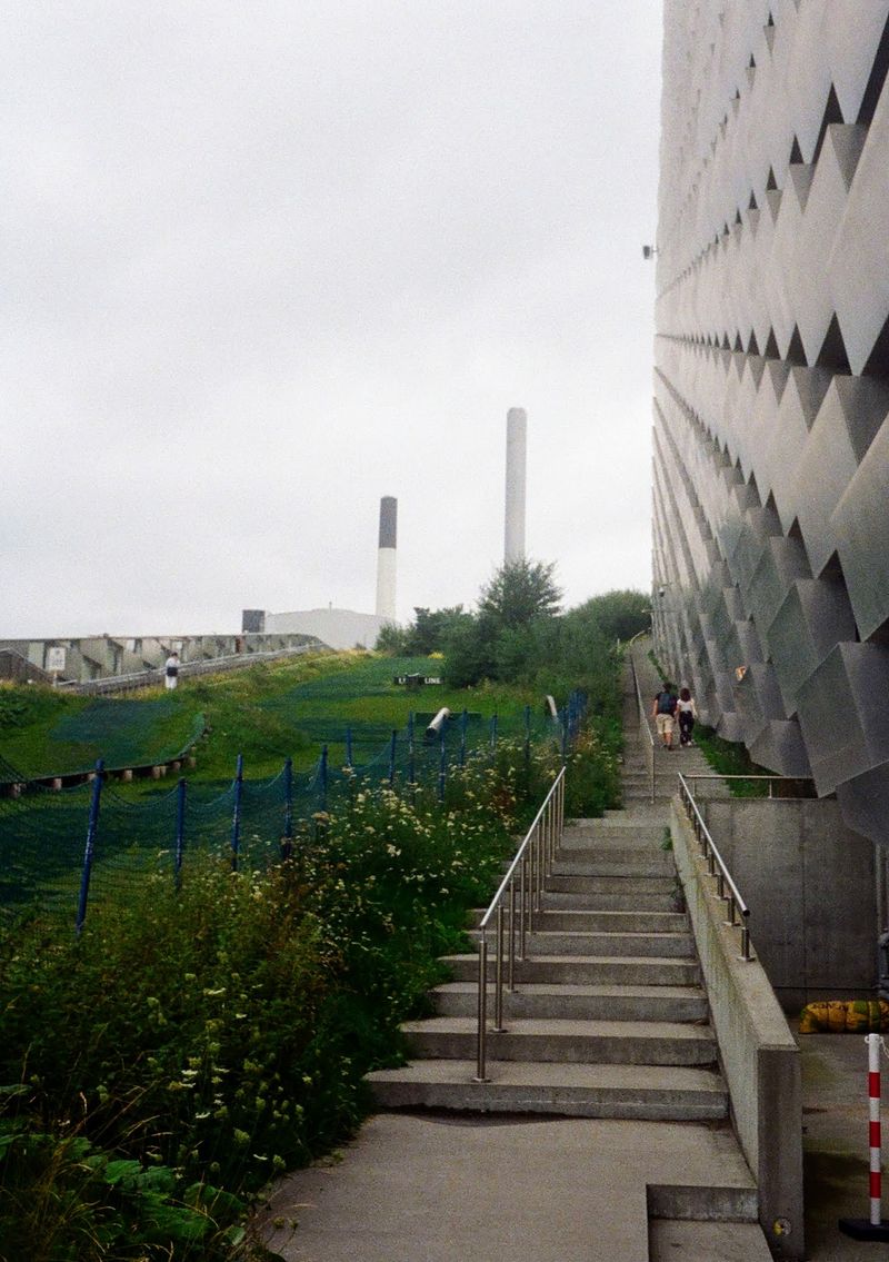 The green slopes of a ski hill run down away from the smoke stacks of the factory below.