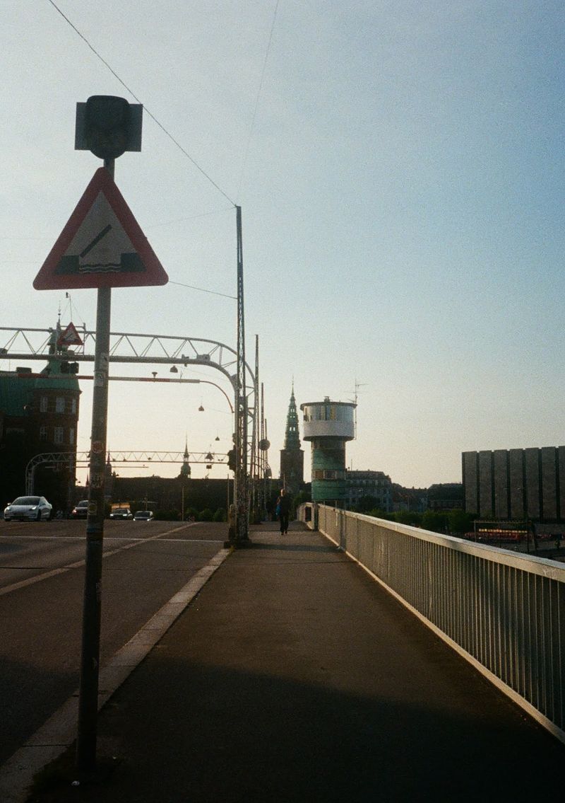 A man walks over a bridge towards the camera. There is a roadway next to him that has some cars on it. A strange round control tower is to his right, just off the edge of the bridge.