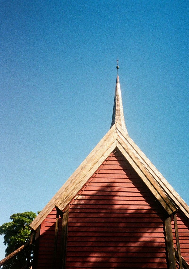 A church spire bathed in sunlight. The red wood of the church building contrasts with the blue sky behind.