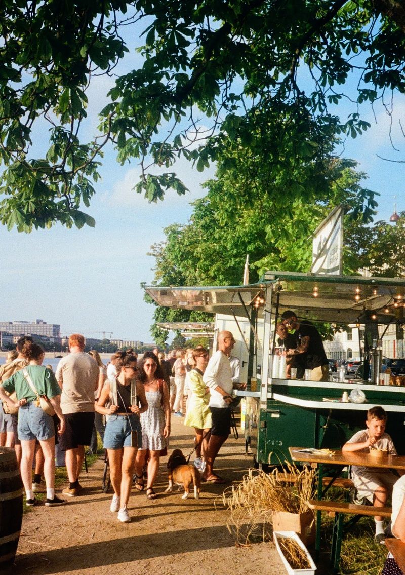 People enjoying the late afternoon sun walk past and line up at food vans that are parked besides the gravel pathway.