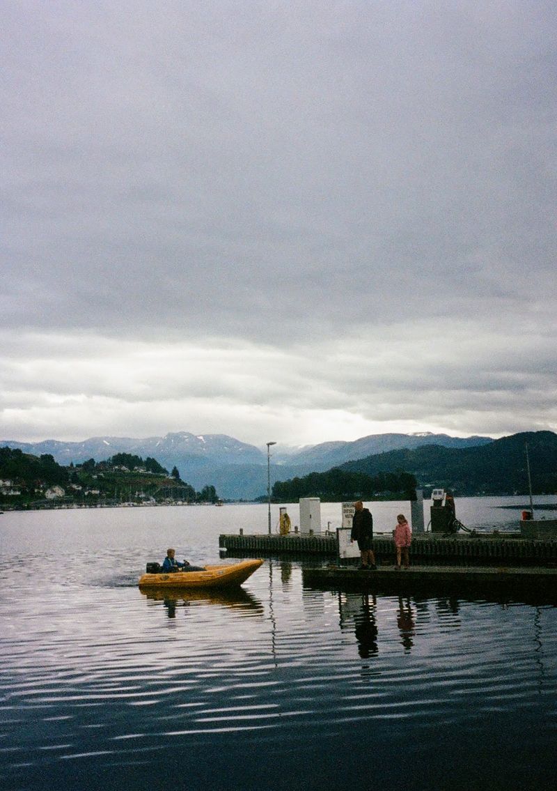 A man stands with a girl on a wooden pier, while a boy carefully navigates a small, yellow, inflatable boat towards them.