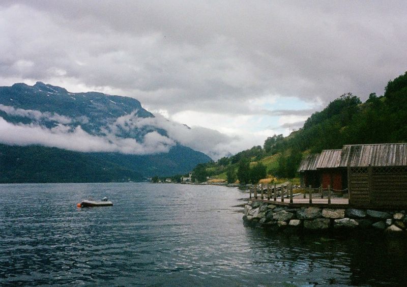 Wooden boat huts sit on the edge of a fjord, with a small boat moored in front of them. The water is calm and the sky is overcast. Mountains in the background have low cloud cover.