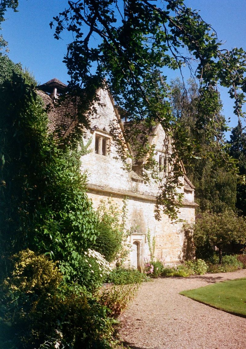 An old stone house sitting at the end of a sandy path. Tree branches overhead frame the shot.