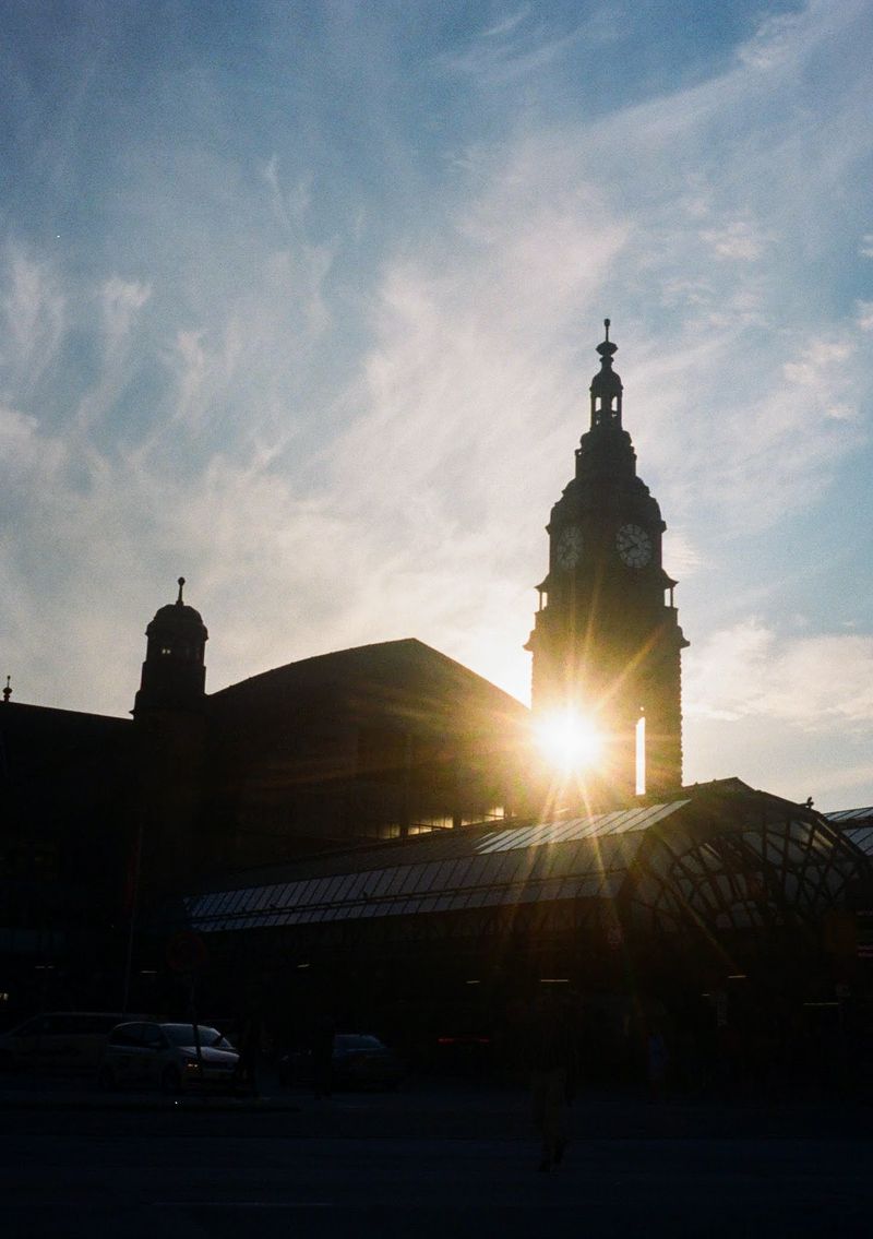 Rays of sunlight shine through a clock tower sitting above a glass covering. Whispy clouds over the blue sky in the background.