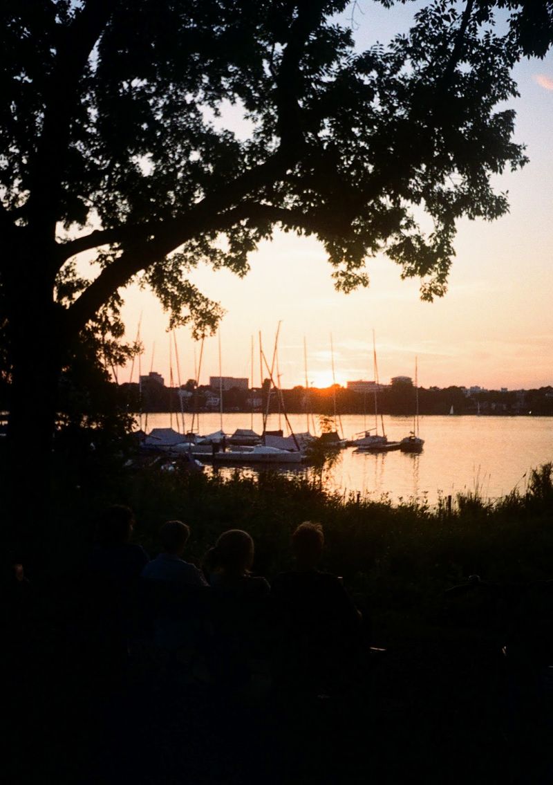 Boats are moared in a lake as the sun sets behind buildings in the distance. In the foreground the shapes of some people can be seen sitting looking out over the lake.