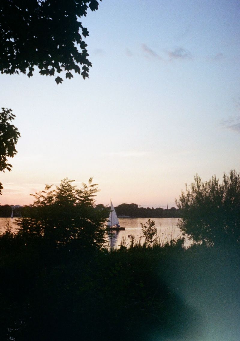 A small single-mast boat sails in a lake at sunset. The lake is lit up in a orange hue, while the sky behind fades from blue to orange. Tree mask the foreground of the shot.